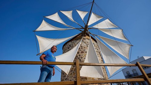 A man leaning against a wooden fence in front of a windmill with white sails and a building in the