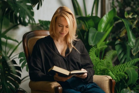 In a serene indoor setting filled with lush green plants, a woman with long blonde hair sits