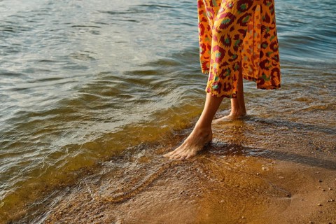 Unrecognizable woman walks barefoot along a sandy beach as waves gently lap at her feet. She wears