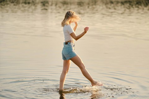 A woman walks through shallow water, splashing lightly as golden sunlight reflects off the surface.