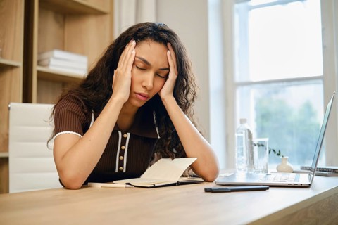 Tired young woman sits at her desk, visibly stressed and holding her head in her hands. The