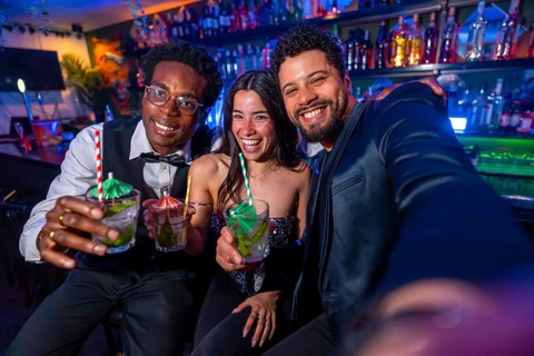 Three friends smiling and capturing a selfie while enjoying colorful cocktails at a lively bar,
