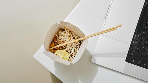 A cardboard bowl of noodles with vegetables and lime sits next to a laptop on a white table. The