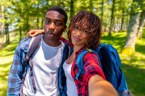 Two young hikers with backpacks smiling while taking a selfie in a sunlit forest, capturing the joy