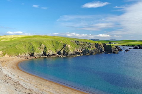 Green hills hug a sandy coastline with calm blue waters and wide open skies, Hillswick, Eshaness,