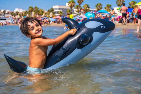 Smiling child having fun riding an inflatable orca toy in the shallow sea water by the beach on a
