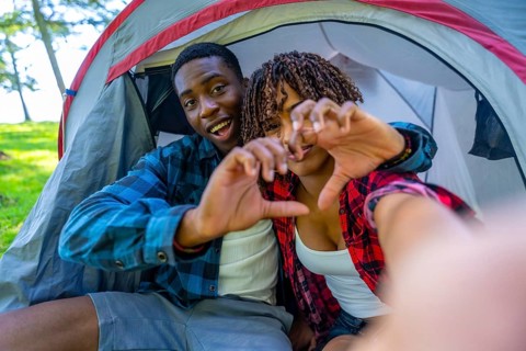 Young smiling couple making heart shape with hands taking selfie inside tent during camping trip
