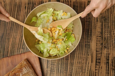 Hands are stirring a bowl of fresh salad featuring crisp lettuce and sweet corn. Nearby, a rustic