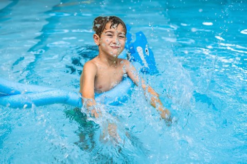 Smiling young boy splashing in a swimming pool, using a blue pool noodle. Joyful summer activity