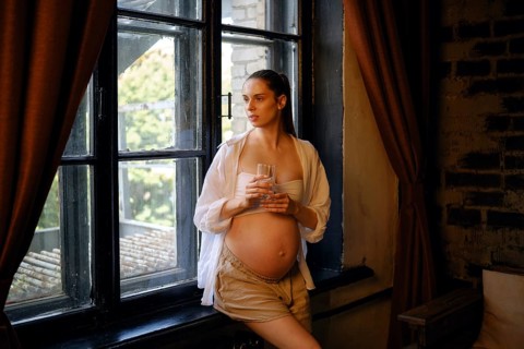 A pregnant woman stands by a large window in her home, holding a glass of water. She gazes outside,