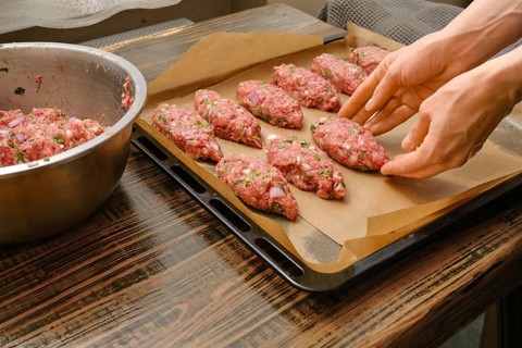 Hands shape meat patties on a lined baking tray in a warm kitchen. This preparation hints at a