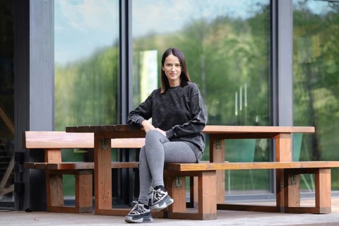 A young woman sits comfortably at a large wooden table placed outdoors. She wears a casual outfit