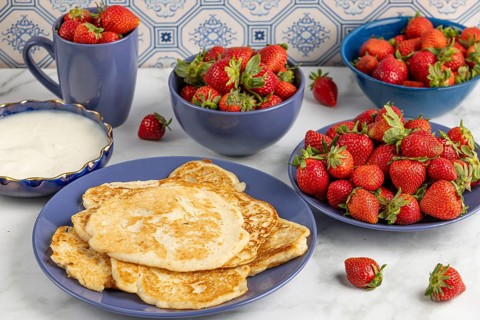 Pancakes on a plate, surrounded by bowls of strawberries and a bowl of yoghurt