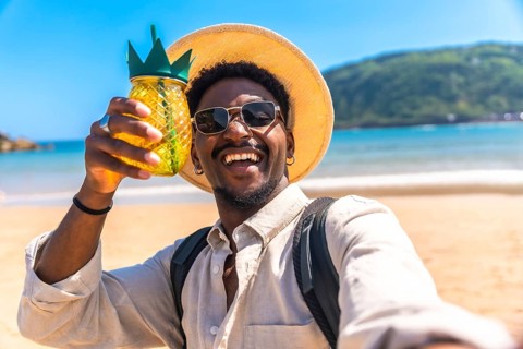 Young man wearing sunglasses and straw hat holding pineapple shaped cocktail while taking selfie on