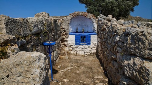 Small stone chapel with blue façade embedded in ancient ruins under a bright blue sky, Artemis
