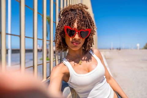 Fashionable young woman with curly hair taking a selfie by the beach, wearing trendy heart shaped
