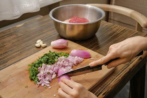 A person chops purple onions and fresh herbs on a wooden cutting board. A bowl of ground meat sits