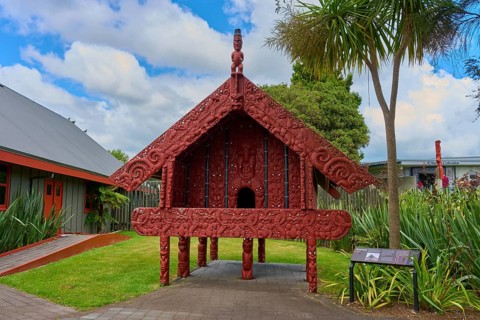 Small Maori building with red carvings surrounded by palm trees, New Zealand Maori Arts and Crafts