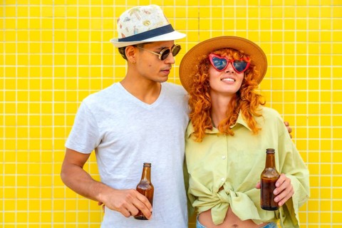 Young couple wearing hats and sunglasses, holding beer bottles and smiling joyfully against a