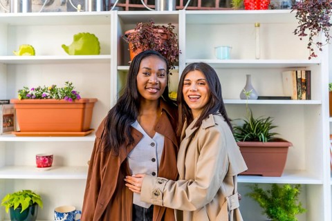 Two happy female friends are smiling and embracing in a plant shop, surrounded by shelves filled