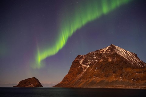 Aurora borealis in green colors over mountain range and coastline at night, Lofoten, Norway
