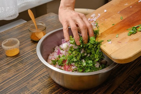 A hand adds freshly chopped cilantro and onions into a large metal bowl. The cozy kitchen