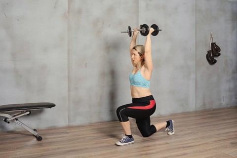 A woman is engaged in a strength training workout, lifting dumbbells overhead while in a lunge