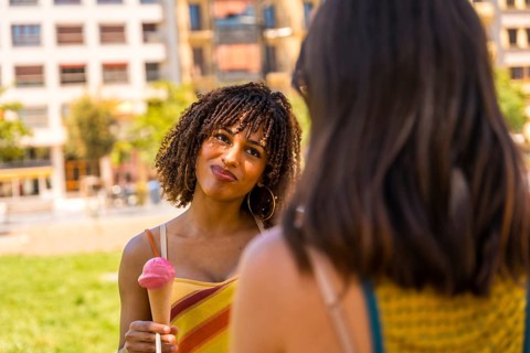 Young woman enjoying a strawberry ice cream cone while talking to her friend in a city park on a