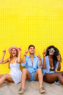 Group of cheerful multi ethnic friends pointing up while sitting on the sand against a vibrant