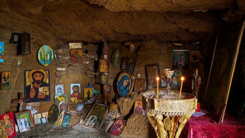 Icon-decorated cave altar with glowing candles, surrounded by religious artworks and paintings,