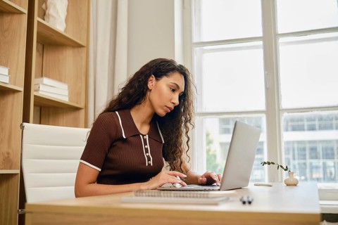 In a cozy home office, a woman with long, curly hair focuses intently on her laptop while seated at