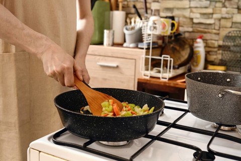 A person stirs a colorful mix of fresh vegetables in a frying pan on a stove in a cozy kitchen.