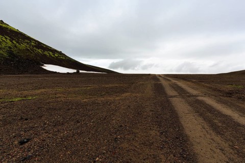 Gravel track, barren volcanic landscape with snowfields, mountain peaks with rain clouds,