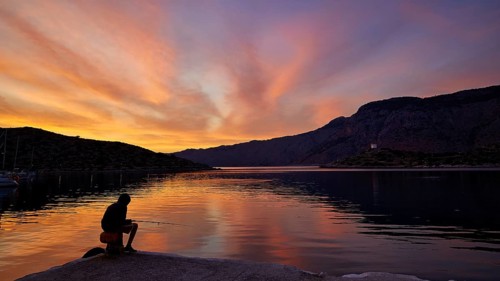 Angler sitting on the lakeshore during a colourful sunset against a mountainous backdrop, Holy