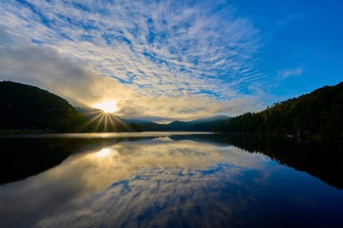 Sunbeams break through clouds and are reflected in a calm lake surrounded by green mountains,