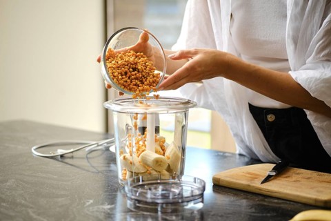 A person carefully adds sea buckthorn berries to a blender containing sliced bananas. The setting