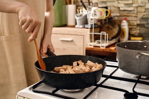 A person stirs diced pork meat in a frying pan on a gas stove. The kitchen is modern, featuring