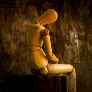 A wooden mannequin is positioned on a rough wooden surface, looking contemplative against a