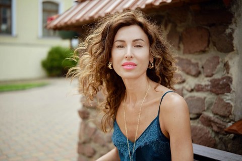 A woman with beautiful curly hair sits on a bench next to a rustic stone wall. She wears a stylish
