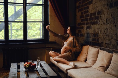 Relaxing at home, a pregnant woman sits on a cozy couch, taking a selfie and posting it in her