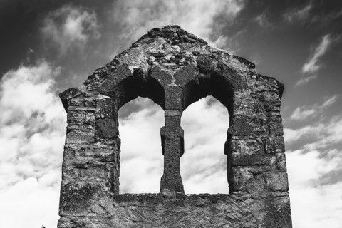 Crumbling stone ruins of a bell tower stand against a cloudy sky, showcasing two arched windows.