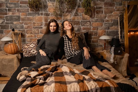 Two women relax together on a bed surrounded by autumn decor, including pumpkins and warm blankets,