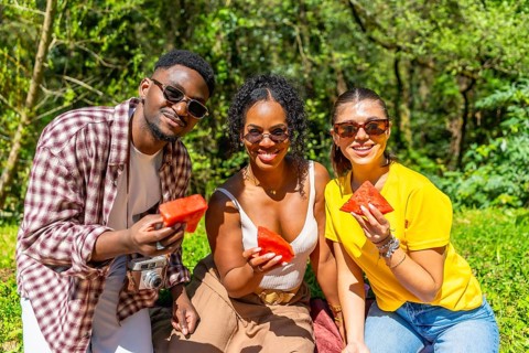 Three friends are sharing watermelon slices during a summer picnic in a lush park, enjoying the