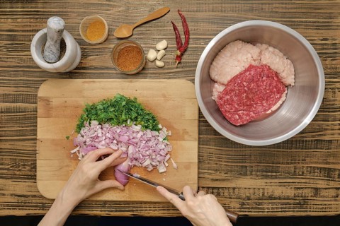 Hands chop shallots and parsley on a wooden cutting board. Ground meat is prepared in a bowl