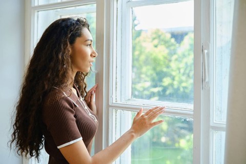 A young woman with long curly hair stands by a window, thoughtfully gazing outside. Sunlight