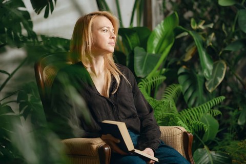 In a serene indoor garden, a woman sits comfortably in a chair holding a book. Soft light filters