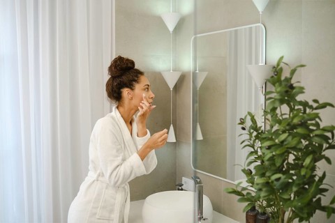 A woman in a white bathrobe stands in front of a mirror, applying skincare products to her face