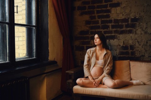 A relaxed pregnant woman sits on a couch in her home, gazing thoughtfully out the window. Soft