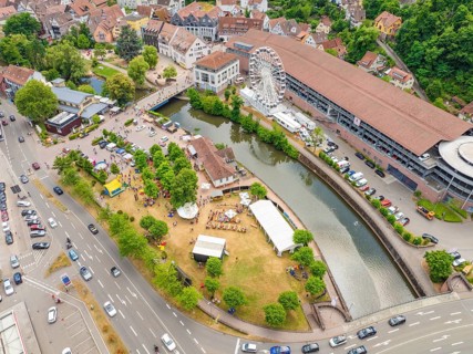 Park with people by a river, flanked by town buildings and a Ferris wheel, with heavy road traffic,