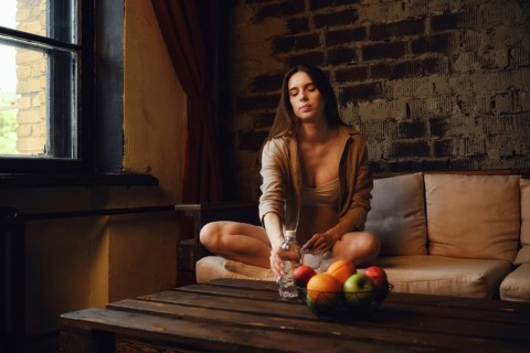 A pregnant woman is sitting on the couch near a window and reaching for a bottle of water on the